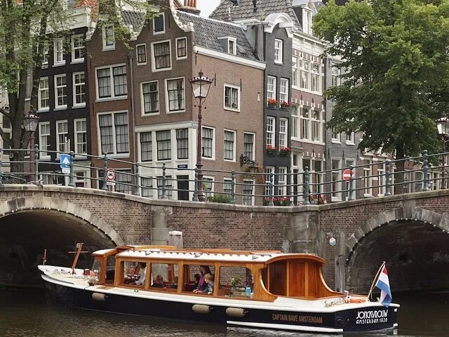 Klassisches Grachtenboot fährt unter einer Amsterdamer Brücke in der Nähe von Häusern hindurch
